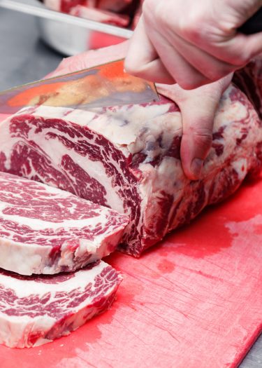 Closeup cook's hand cuts ribeye marbled beef steak with sharp knife on red plastic cutting board on metal table in restaurant kitchen. Concept steakhouse specializing in grilled meat, live fire